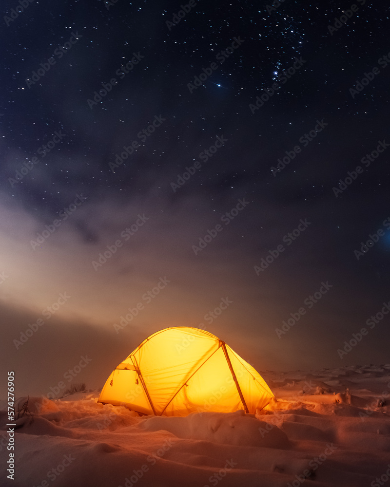 Yellow tent lighted from the inside against the backdrop of glowing city lights and incredible starry sky. Amazing night landscape. Tourists camp in winter mountains. Travel concept