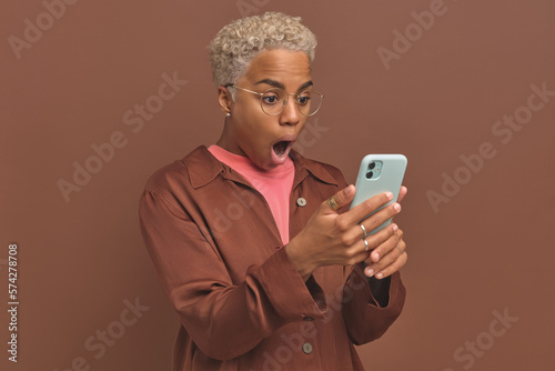 Young shocked beautiful African American woman using mobile phone is dumbfounded by indecent message or photo sent in messenger dressed in casual style posing on isolated brown background. photo