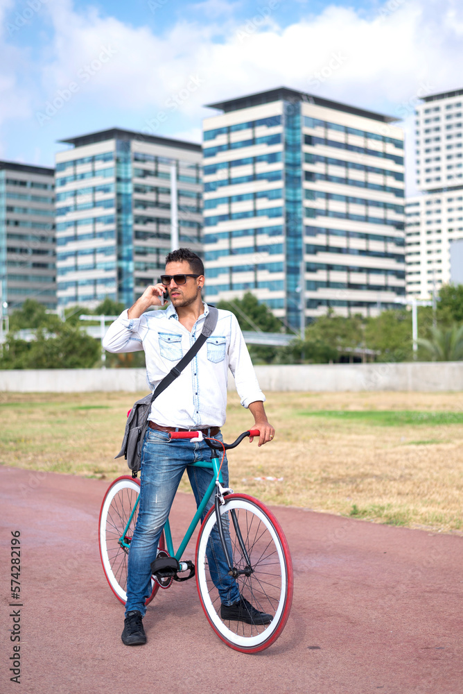 Obraz premium Man with sunglasses standing with his bicycle talking by phone and the city in the background