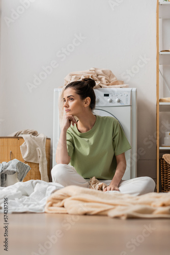 Sad woman sitting near clothes on floor in laundry room.