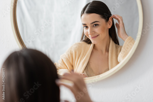 young brunette woman adjusting hair and smiling while looking at mirror in bathroom Fototapet