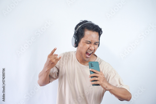 Energized man sings to music, moves actively, wears headphones and casual t shirt, poses against white background, keeps mouth widely opened, enjoys life, isolated on white studio wall