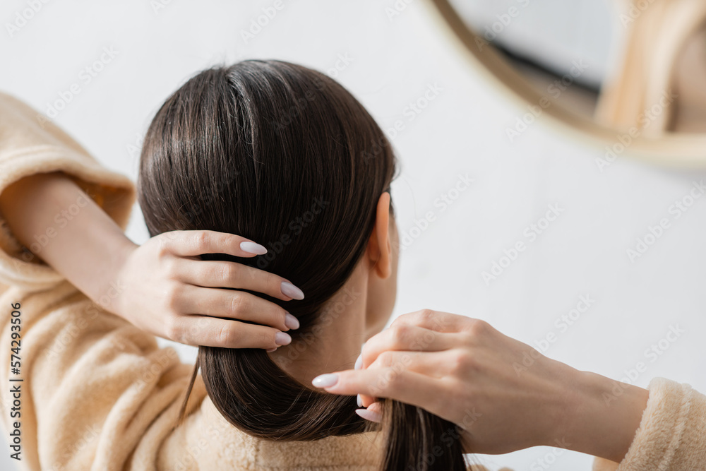 Naklejka premium back view of young brunette woman adjusting shiny hair near mirror in bathroom.