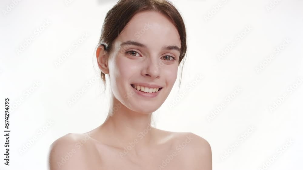 Close-up shot of gorgeous young slim Caucasian woman with long brown hair strokes her shoulder turning her body and smiles widely for the camera on white background | Body care cosmetics commercial Close-up shot of gorgeous young slim Caucasian woman with long brown hair strokes her shoulder turning her body and smiles widely for the camera on white background | Body care cosmetics commercial