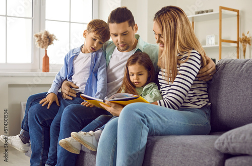 Young family reading book sitting on sofa in living room. Mom, dad, son and daughter enjoying book reading or looking photos in family album. Happy family having good time together at home
