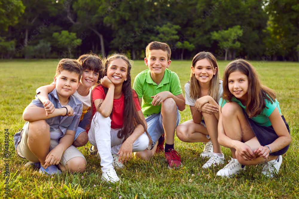 Group portrait of happy children in park. Happy school friends play in nature and enjoy summer together. Six cheerful healthy little Caucasian kids sitting on green lawn, looking at camera and smiling