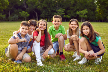 Group portrait of happy children in park. Happy school friends play in nature and enjoy summer together. Six cheerful healthy little Caucasian kids sitting on green lawn, looking at camera and smiling