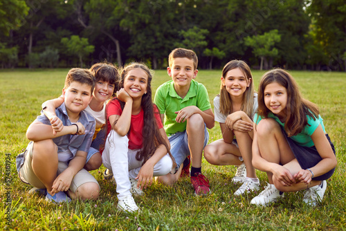 Group portrait of happy children in park. Happy school friends play in nature and enjoy summer together. Six cheerful healthy little Caucasian kids sitting on green lawn, looking at camera and smiling © Studio Romantic