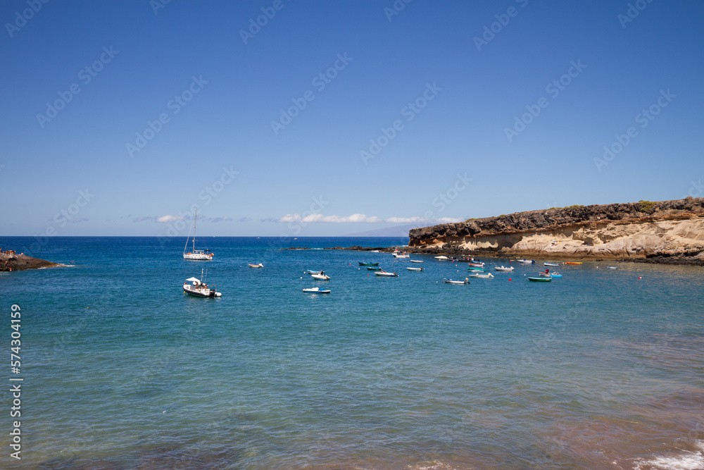 Bay views of a small fishing village with boats at anchor in a calm turquoise ocean with yellow hills in the background. La Caleta, Tenerife, Canary Islands Spain.