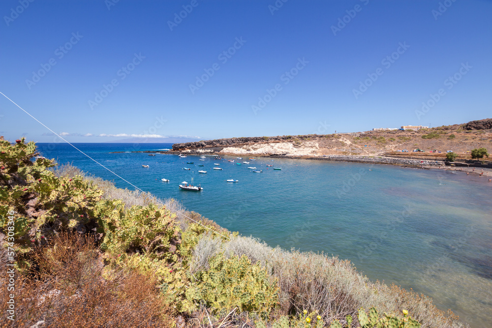 Obraz premium Bay views of a small fishing village with boats moored in the turquoise calm ocean. Desert plants in foreground and yellow hills in background. La Caleta, Tenerife, Canary Islands Spain.