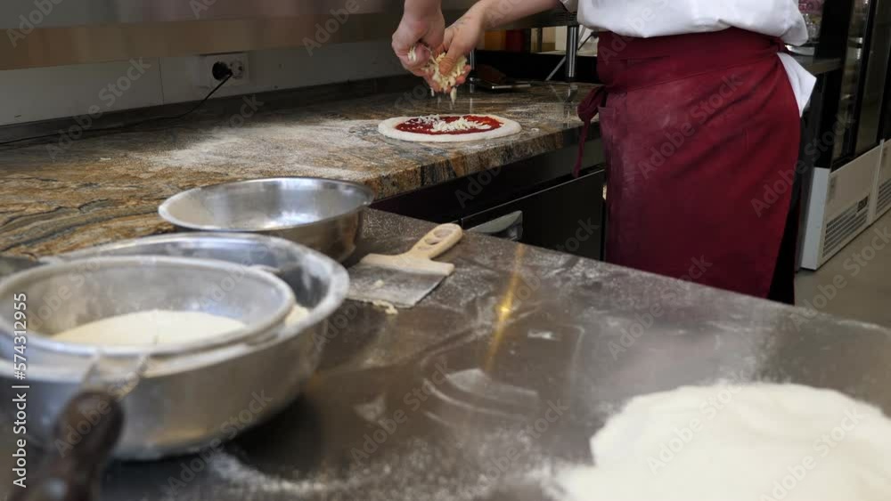 Close-up of the chef pouring grated cheese on the dough with tomato sauce, the process of making pizza in a traditional Italian pizzeria. Close-up of the chef pouring grated cheese on the dough with tomato sauce, the process of making pizza in a traditional Italian pizzeria.
