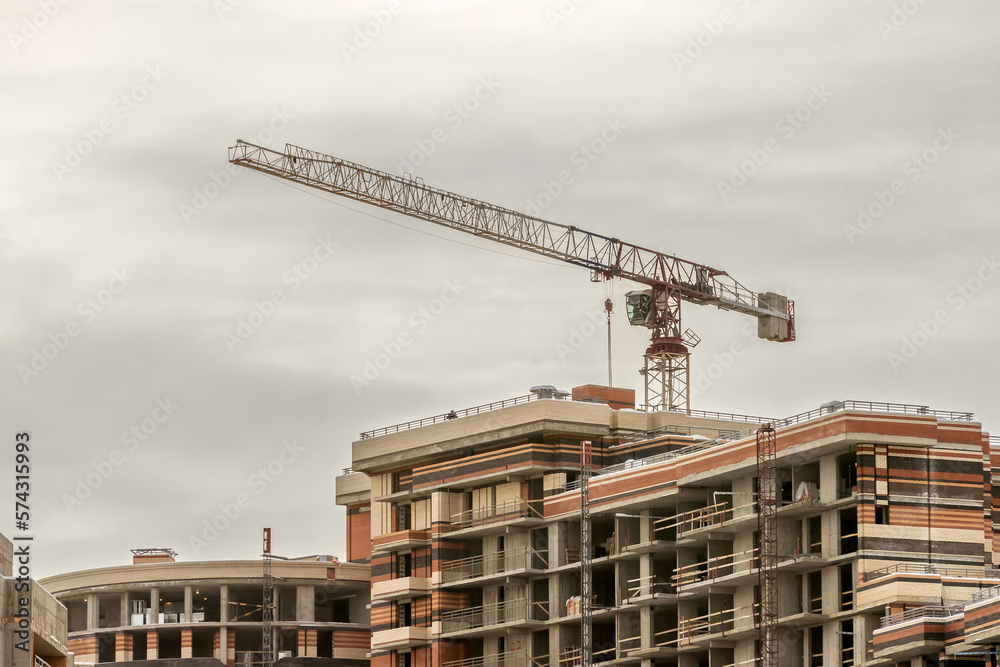 A construction crane against the background of a sky with gray clouds and a multi-apartment building under construction