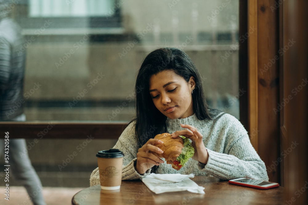 A young African American woman sits in a cafe and eats an appetizing burger. Bad cheap food. Wrong nutrition.