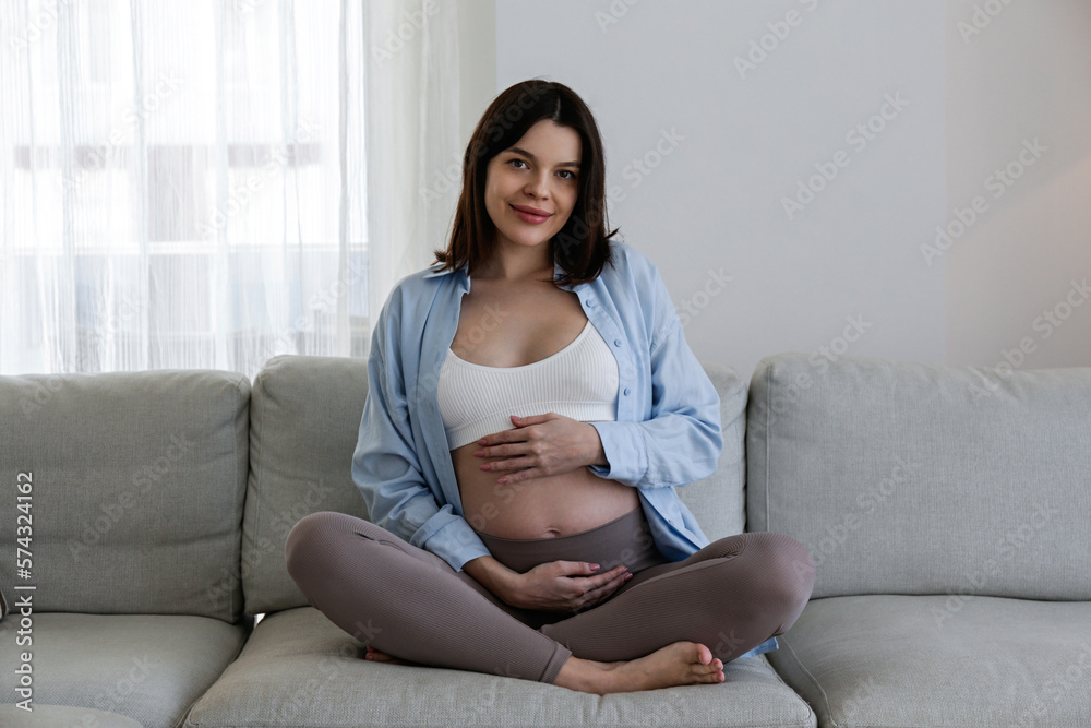 Brunette young woman on second trimester of pregnancy. Pregnant female sitting on the couch with arms on her belly. Expecting a child concept. Background, copy space. Brunette young woman on second trimester of pregnancy. Pregnant female sitting on the couch with arms on her belly. Expecting a child concept. Background, copy space.