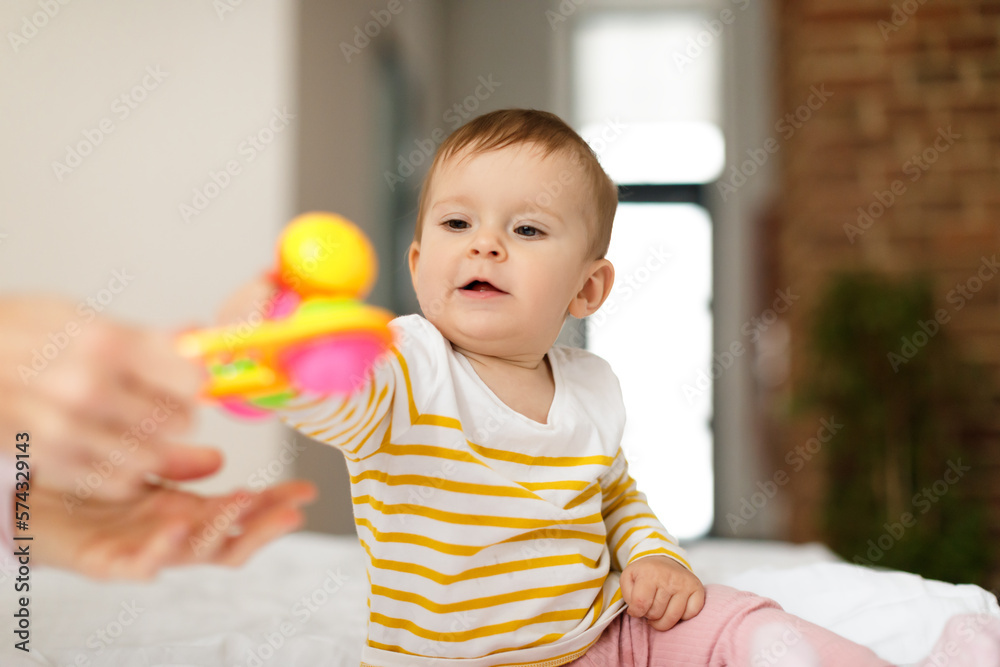 Cute baby girl taking rattle toy from mother hands, sitting on bed, little child in bodysuit resting at home with mom