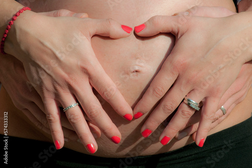 Female and male hands are intertwined on the belly of a pregnant woman  forming a heart with their fingers