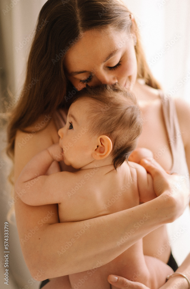 The portrait of young brunette mom kissing her sweet baby girl who is with her finger into the mouth; mother's day concept The portrait of young brunette mom kissing her sweet baby girl who is with her finger into the mouth; mother's day concept