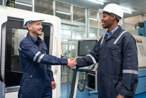 Two male engineer shaking hands after work success at factory. Smiling technician wearing uniform and helmet safety operator CNC machine in workshop heavy metal industrial