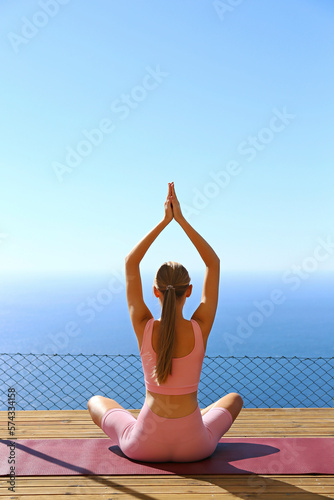 Young woman meditating on a terrace with a breathtaking ocean view. Yogini sitting in namaste pose with her palms pressed together above her head. Close up, copy space, background.