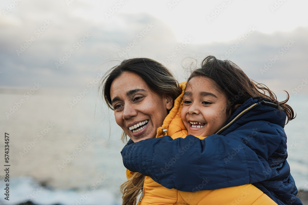 Happy latin mother and daughter having fun on the beach during winter time - Mom and child outdoor