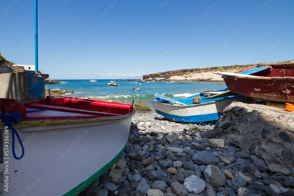 Cheap boats on a pebble beach. Red and white boat on the left, blue and white boat on the right. Bay of turquoise waters and brown and yellow hills in the background. La Caleta, Tenerife, Canary Islan