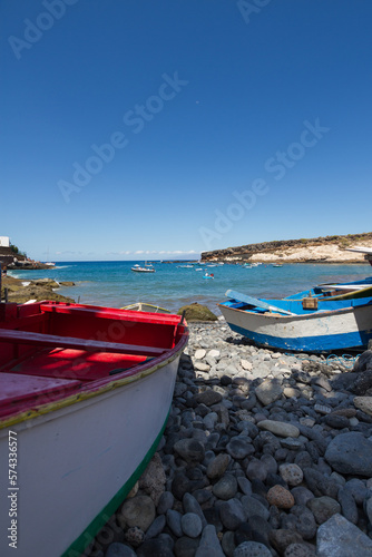 Cheap boats on a pebble beach. Red and white boat on the left  blue and white boat on the right. Bay of turquoise waters and brown and yellow hills in the background. La Caleta  Tenerife  Canary Islan
