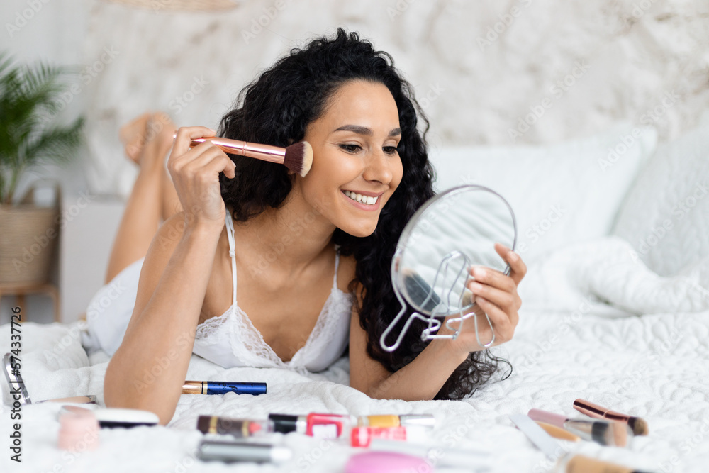 Glad millennial caucasian brunette lady with long hair lies on soft bed applies powder with brush on face Glad millennial caucasian brunette lady with long hair lies on soft bed applies powder with brush on face