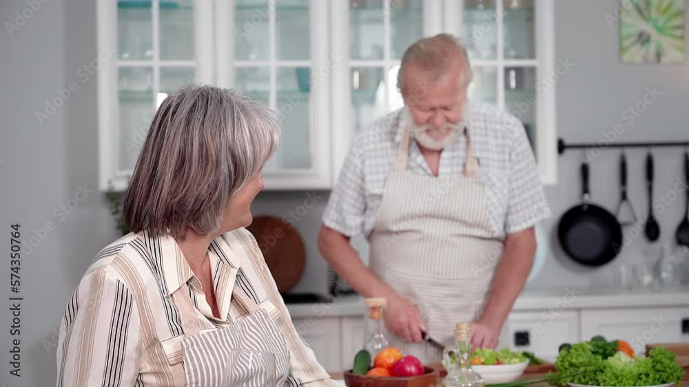portrait of elderly charming woman with glass of wine enjoys her vacation, caring man prepares a romantic dinner in kitchen
