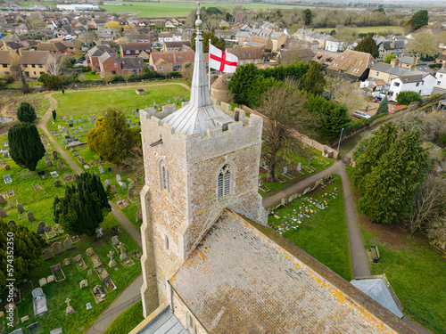 Aerial view of a typical English church showing the St Georges flag flying atop the spire. A cemetery surrounds the village church.