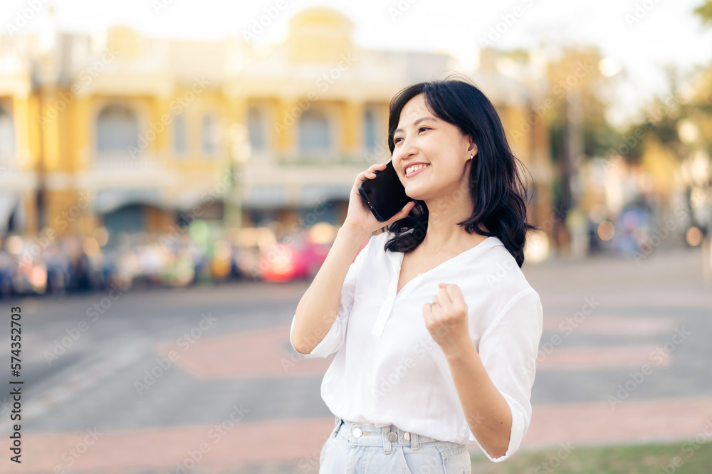 Portrait beautiful young asian woman receiving good news on mobile phone and arms raised feeling cheerful ,happiness. around outdoor street view in a summer day Portrait beautiful young asian woman receiving good news on mobile phone and arms raised feeling cheerful ,happiness. around outdoor street view in a summer day