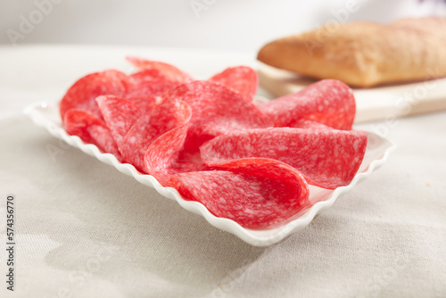 salami slices on a white plate with bread in the background. sausage close-up. meat delicacy on a sunny day. traditional mediterranean snack on a light surface. dried sausage serving. antipasta macro