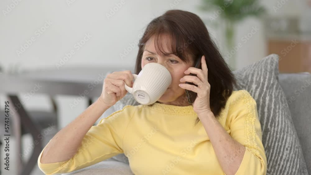 Beautiful mature asian woman long hair wearing yellow top talking on mobile phone smiling drinking coffee sitting at home living room table blurred plant background Beautiful mature asian woman long hair wearing yellow top talking on mobile phone smiling drinking coffee sitting at home living room table blurred plant background