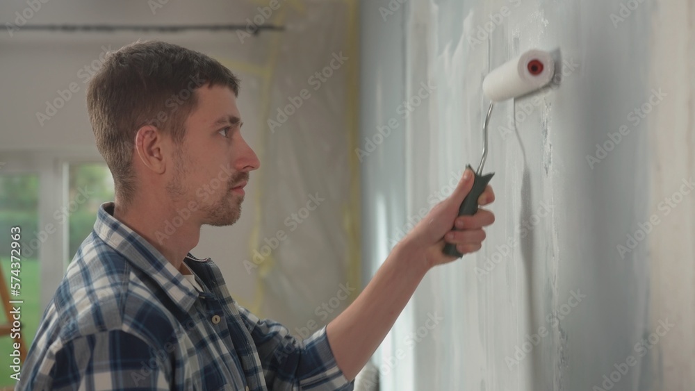 Side view of a portrait of a young man is painting wall with white paint using a paint roller. Male painter in checkered shirt enjoying renovations in the apartment. Concept of repair. Side view of a portrait of a young man is painting wall with white paint using a paint roller. Male painter in checkered shirt enjoying renovations in the apartment. Concept of repair.