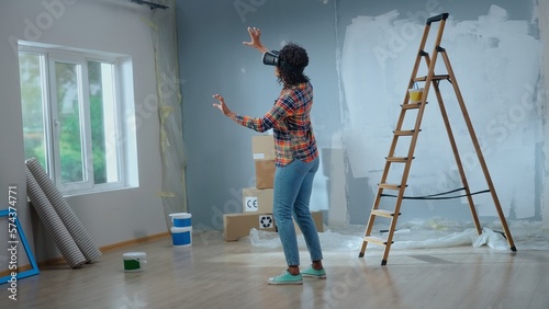 African american woman in virtual reality headset furnishes interior of room. Female makes choice of wallpaper, furniture, curtains using virtual technology. Concept of modern apartment renovation.