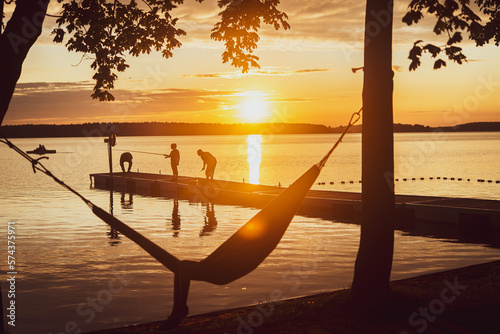 Summer sunset by the calm lake with people relaxing on jetty,  fishing, lying on hammoc, copy space photo