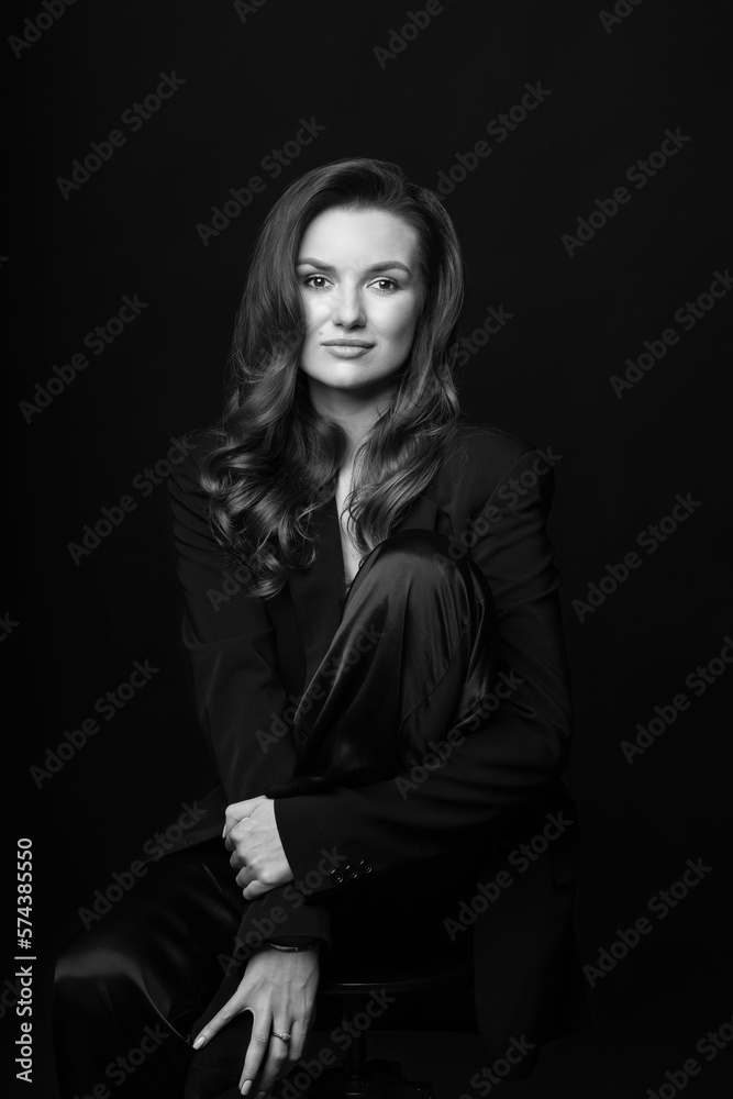 Portrait of beautiful woman with black suit and big wavy hair in dark studio background. Model sitting on chair and hugging her one leg. Woman looking at camera. Black and white image