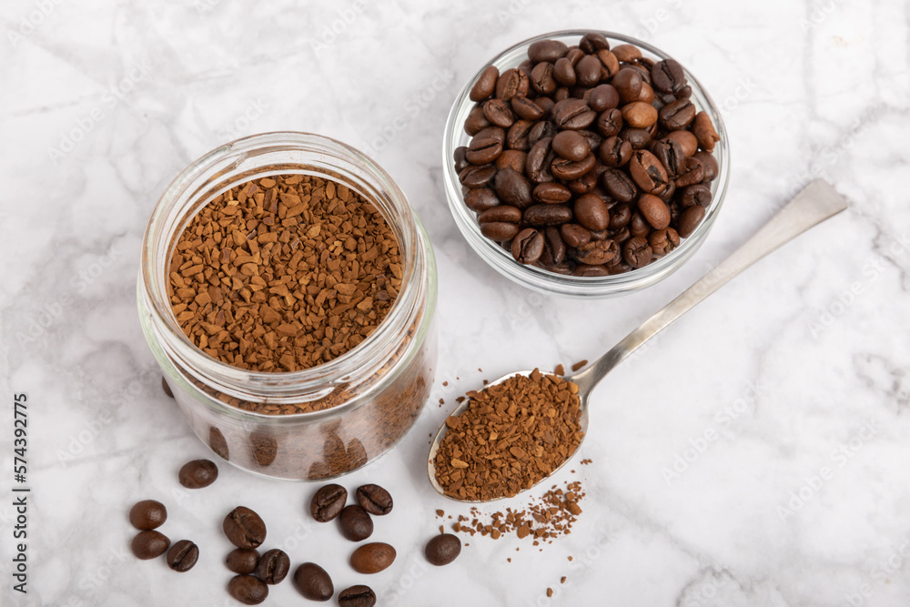 Instant coffee beans in a glass jar on a white marble background. Hot drink ingredients. Instant coffee on the table. Espresso. Place for text. Place to copy. Banner. Instant coffee beans in a glass jar on a white marble background. Hot drink ingredients. Instant coffee on the table. Espresso. Place for text. Place to copy. Banner.