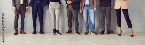 Legs of unrecognizable business people standing in a row on a grey office wall bacground. Cropped shot of a stylish group of employees in casual clothes waiting for job interview. Banner.