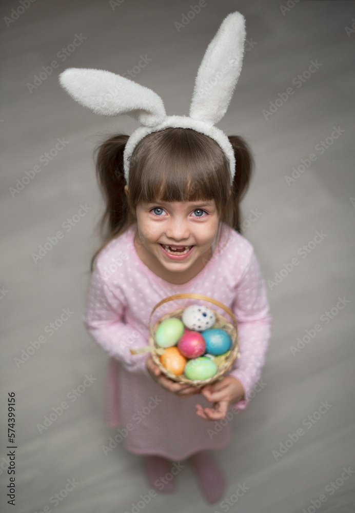A portrait funny little girl with brown hair and a toothless smile, wearing bunny ears and holding basket of Easter eggs in her hands. Soft focus. Copy space. 