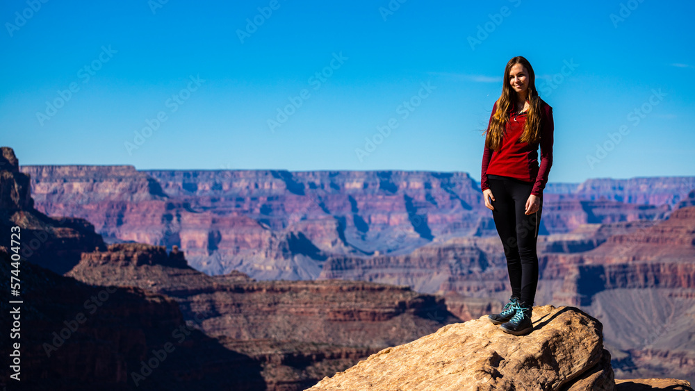 Naklejka premium Beautiful long-haired smiling tourist girl stands with her back to the Grand Canyon. Traveling in winter in Grand Canyon National Park, Arizona, USA.