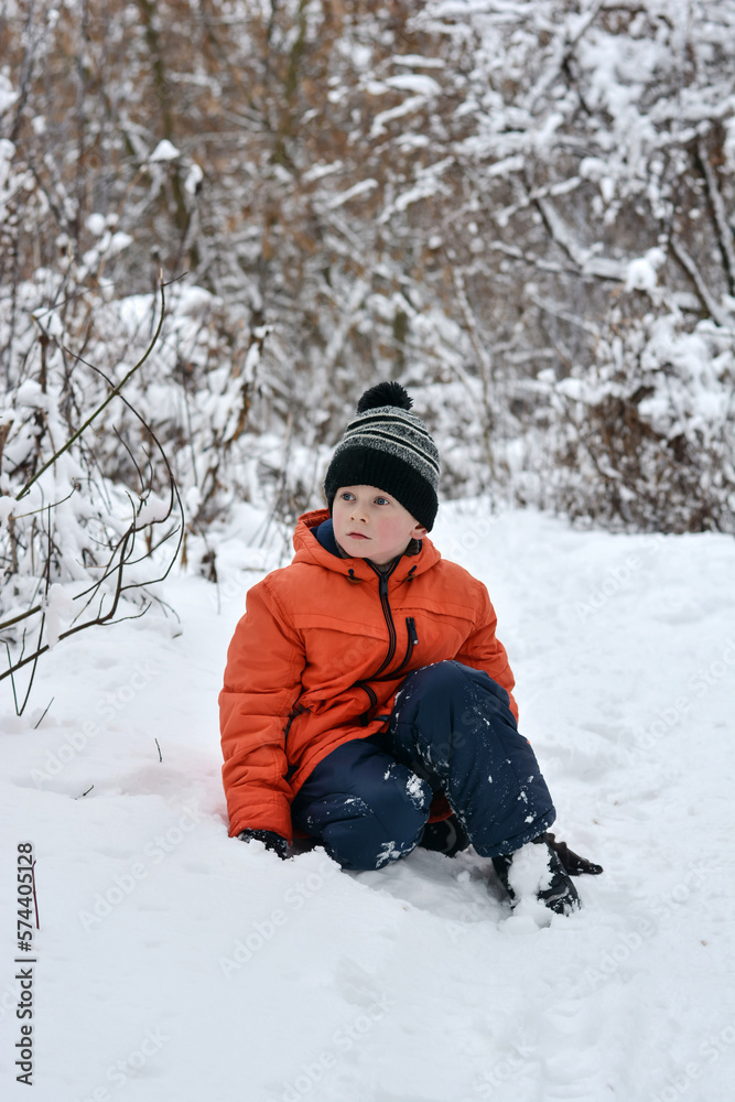 A boy in a jacket and a hat sits in the snow and looks into the distance with surprise. Winter vacation.