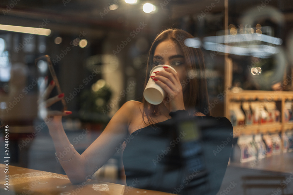 Pretty young woman in a black top drinking coffee, reading the news feed on smartphone and sitting by the window in a cafe. Lifestyle girl