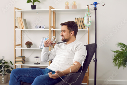 Dehydrated young man receiving intravenous vitamin therapy in hospital room. Male patient sitting in armchair attached to vitamin IV infusion drip in wellness center or beauty salon photo