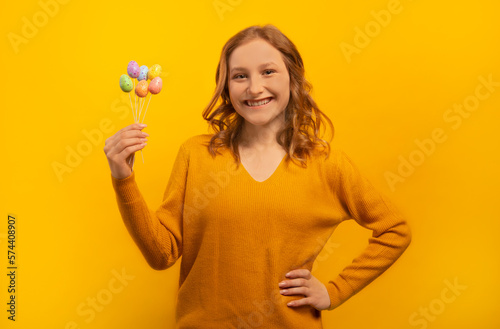 Happy smiling laughing young woman in yellow sweater holding small painted dyed easter eggs on sticks, keeping hand on waist hip isolated on yellow background.Easter Day celebration concept.