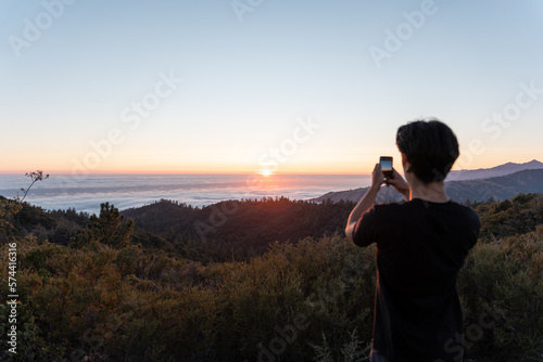 A young man is taking a photo of the sunset landscape with his phone