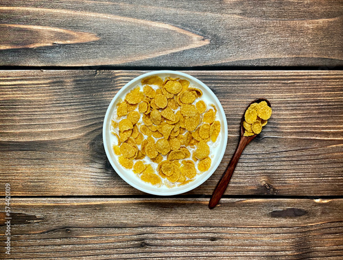 Corn flakes with milk in white plate with wooden spoon on wooden background. Top view, flat lay.
