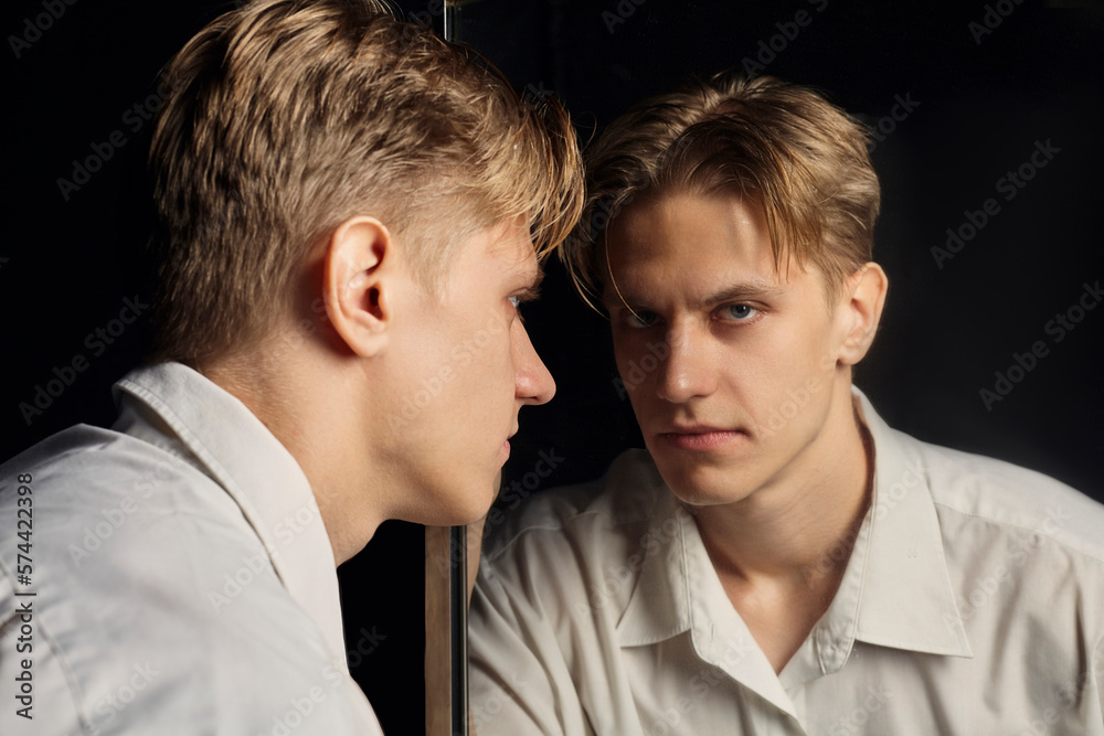 a serious man in a white shirt looks at himself in the mirror standing on a black background