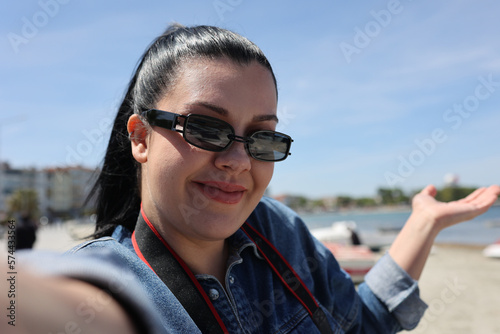 Smiling girl taking selfie with mobile phone by sea. She has sunglasses.