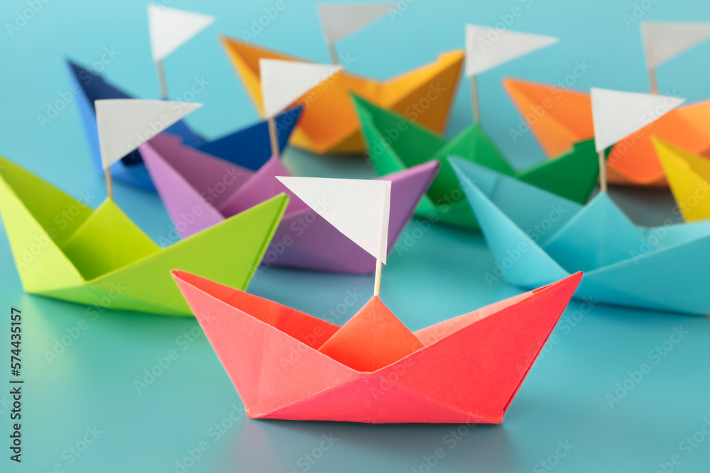 Colorful paper ships on blue background with white flags indicate direction, unity, competition, goals concept. Colorful paper ships on blue background with white flags indicate direction, unity, competition, goals concept.
