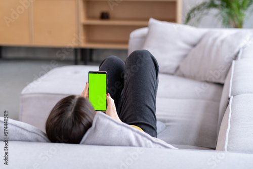 Feminine Hand Scrolling Feed on Smartphone with Green Screen Mock Up Display. Female is Relaxing on Sofa at Home, Watching Videos and Reading Social Media Posts on Mobile Device. © Inna